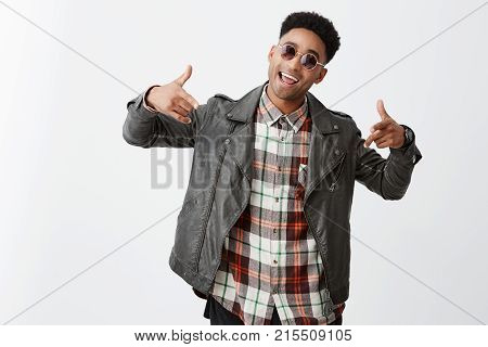Wazzup man. Attractive young dark-skinned man with afro haircut in black leather jacket and sun-glasses laughing, gesticulating with hands, dancing and singing on party, having fun