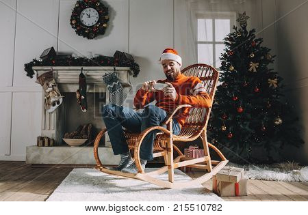 Adult and bearded guy is sitting in the rocking chair in a beautiful white room decorated with Christmas toys, tree and garlands. He is watching a Christmas special series of his favourite program on the phone. Guy is happy and full of joy.