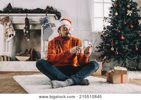 Another picture of a man sitting on the carpet with his legs crossed in a white Christmas decorated room and chatting with someone on the phone. He is also ordering some good yummy food for dinner when his friends will come.