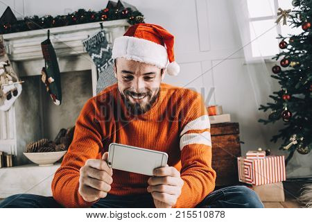 Close up of adult and cheerful guy sitting on the floor in a white Christams decorated room and looking to the phone. He is holding it in his hands and smiling because he os talking with his parents on Skype. Cut view