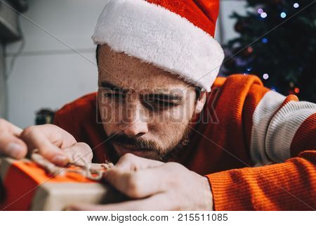 Close up of a bearded guy in red sweater packing a gift for his friend in a white room near the Christmas tree. He is doing that neatlywith full concentration. Young man hopes his friend will like the present. Cut view