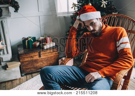 Close up of a guy in red hat and sweater sitting in the rocking chair in a bright and white room decorated in Christmas style. Young man is bored and sad. He doesn't have a Christmas mood at all. Cut view