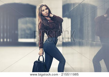 Happy young woman with handbag in city street. Stylish fashion model in leather fringe suede jacket and blue jeans
