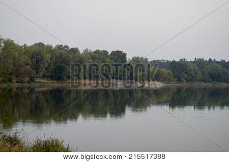 Wide river flowing across green forest. Fall. Evening. Reflections of trees in the calm water. Sundown
