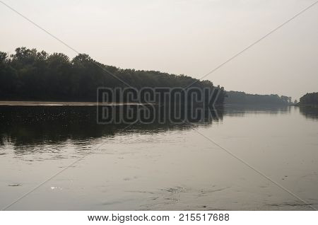 Wide river flowing across green forest. Fall. Evening. Reflections of trees in the calm water. Sundown