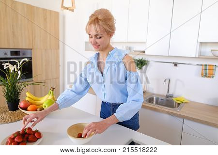 Slender, sweet woman cooks delicious breakfast of oatmeal and ripe red strawberry, with smile on face, takes plate in hands and brings it to camera lens, standing in stylish and light kitchen with white cabinets, household appliances and variety of utensi