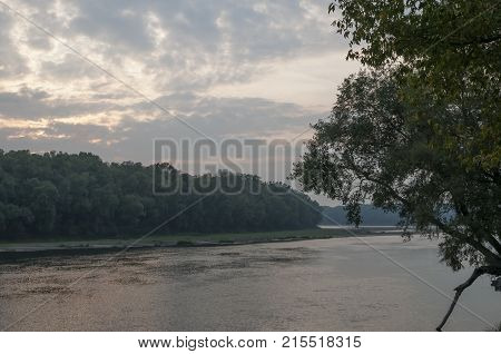 Wide river flowing across green forest. Fall. Evening. Reflections of trees in the calm water. Sundown