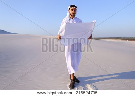 Beautiful Emirate Sheikh businessman studying project of future construction of complex, standing in middle of bottomless desert with white clean sand on clear warm day against blue sky. Swarthy Muslim with short dark hair dressed in kandura, long, spacio