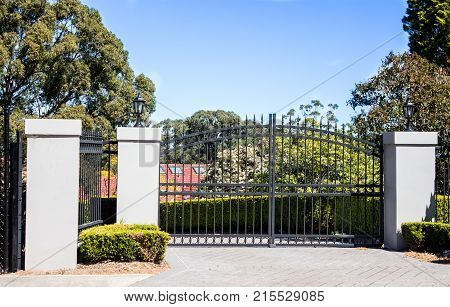 Black metal driveway entrance gates set in brick fence with garden trees in background