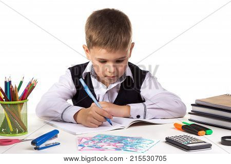 Cute school boy sitting at the desk, writing, surrounded with stationery.