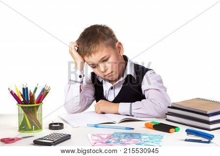Puzzled bored pupil sitting at the desk with hand under the head surrounded with stationery.