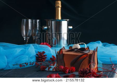 Sweet appetizing beautiful chocolate cake with two crystal glasses of champagne and cooler for champagne on dark background. Cake with blue chiffone for birthday party
