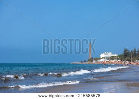 Vacation at the seaside: coastal view on the Maspalomas beach with people and the old Lighthouse in the background. Maspalomas, Grand Canary, Spain - July 18 2017.