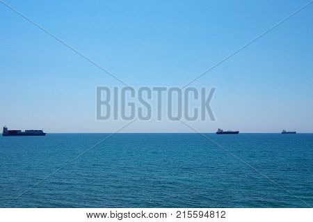The blue calm sea. Not big waves. A clear sunny day. A blue sky shining towards the horizon. By the sea there is a caravan of three ships. Barge ships are carrying cargo.