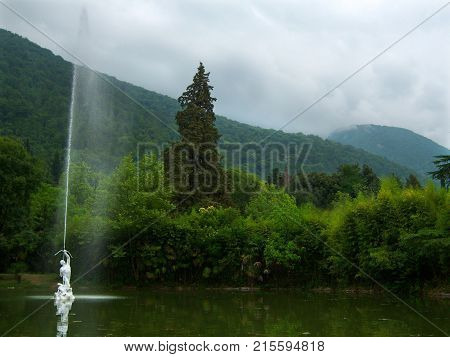 Pond with green water. In the center of the pond a fountain in the form of a man shooting from a bow upwards. A stream of water escapes from the arrow. The pond is arranged in an old overgrown park. Around the trees. In the background mountains are overgr