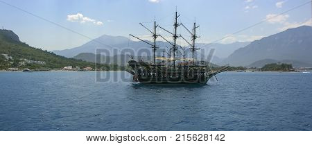 A pirate ship at the dock. Brick near the shore. The schooner in the turquoise sea. Sailing ship in the sea against the backdrop of the mountains. Tourist boat for walks. A ship with sails lowered.