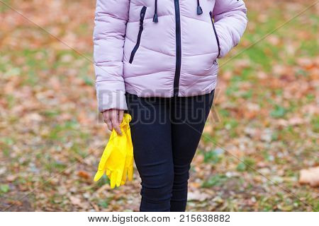 Close-up of a girl in an autumn park, outdoors, stands with rubber, yellow gloves in her hands, to harvest the fallen leaves.