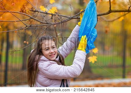 A girl in yellow, rubber gloves, stretches to a branch on a tree to remove the package that is stuck there, looks into the camera.