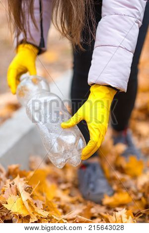Close-up of a girl's hand in yellow, rubber gloves with a plastic bottle on the background of fallen autumn leaves.
