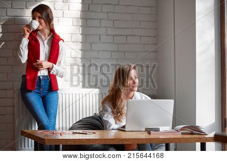 Beautiful young woman smiling joyfully thinking while working on her laptop her female friend classmate drinking coffee on the background copyspace students educational learning lifestyle concept.