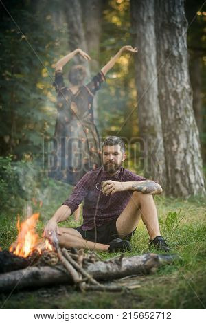 Man and woman relax at bonfire in forest. Girl dance and hipster drink tea at campfire. Camping hiking recreation lifestyle. Summer vacation concept.