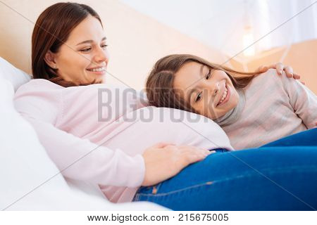 Waiting for answer. Adorable attractive girl  leaning to the tummy  while beautiful woman embracing her with a hand and looking at her