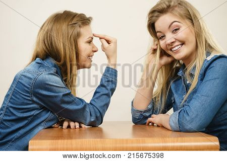 Woman saying bad things about her over confient female friend. Pointing at forehead stupidity gesture.