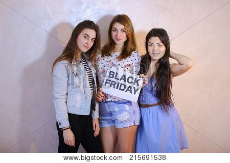 Lovely girls with smiles on their faces look at camera and pose, hold sign in their hands denoting with Black Friday texts, discounts and purchases, stand in room against background of light wall. European-looking girl with red hair dressed in bright T-sh