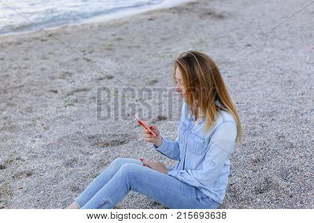 Cute girl holding cell phone and leads in social network or post photos, laughing and smiling, sharing impressions of beauty of sea, sitting on sandy beach on backdrop of sea and sky on clear evening . Young European-looking woman with long blond hair dre
