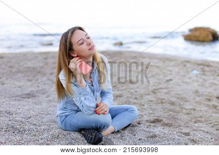Cute girl holding cell phone and leads in social network or post photos, laughing and smiling, sharing impressions of beauty of sea, sitting on sandy beach on backdrop of sea and sky on clear evening . Young European-looking woman with long blond hair dre
