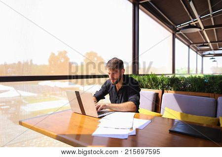 Prosperous financial manager enjoying new project idea and working with laptop and documents. Attractive successful man wears black shirt and watch, sitting at cafe table. Concept of clear budgetary planning, providing financial guidance and support to cl