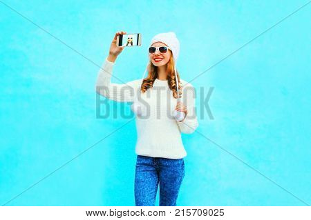 Woman Takes A Picture Self Portrait In White Knitted Sweater, Hat On A Smartphone On Blue Background
