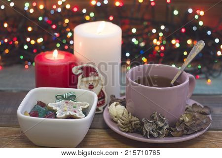 Festive still life with a pink porcelian cup of tea, a saucer, a teaspoon, bowl of colored candies, two burning candles and fairylights on the back, a colored wooden background