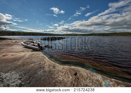 Empty white inflatable boat with motor on the rocky lake shore. Sun flare or boche effects in the left side of the sky and on the rock in the right lower corner of the photo.