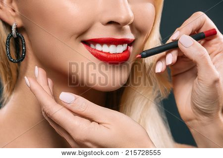 cropped image of beautiful blonde hair girl with visagiste applying lipstick with brush isolated on black