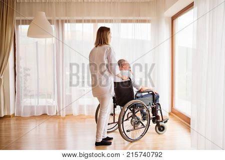 Health visitor or a nurse and a senior man in a wheelchair during home visit. Man and woman looking out of a window.