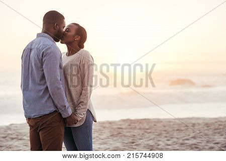 Romantic young African couple standing together on a sandy beach at sunset holding hands and kissing each other