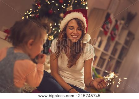 Beautiful young mother and daughter sitting on the floor next to a Christmas tree playing with sparklers.