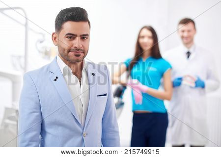 Handsome young man smiling to the camera posing at the dental clinic his dentist and nurse standing on the background copyspace professionalism trust happiness service dentistry oral smile.