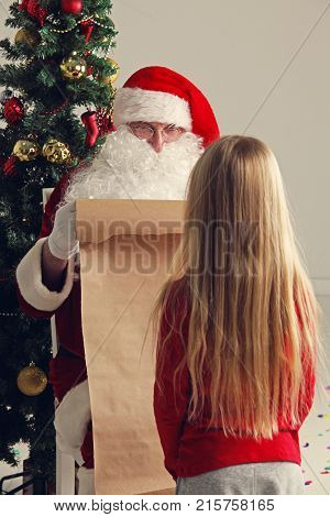 Santa Claus with with list or nice or naugthy list paper with small girl near christmas tree