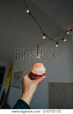 POV of female hand holding pink red velvet cupcake with sparkle candle burning concept burthday or romantic surprise for anniversary or date christmas lights