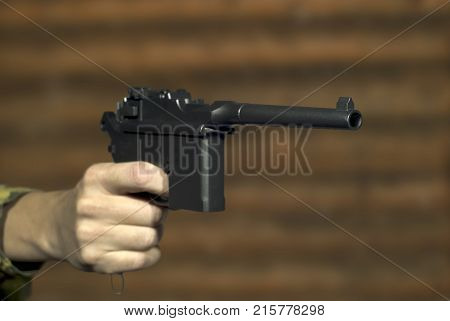 A man's hand with a vintage semi-automatic pistol points past the frame on the background of a log wall. Focus on the muzzle of the pistol the rest is blurry.