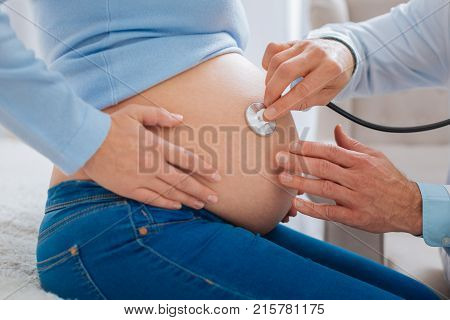 Baby bump. Competent doctor using stethoscope and touching tummy while examining his patient