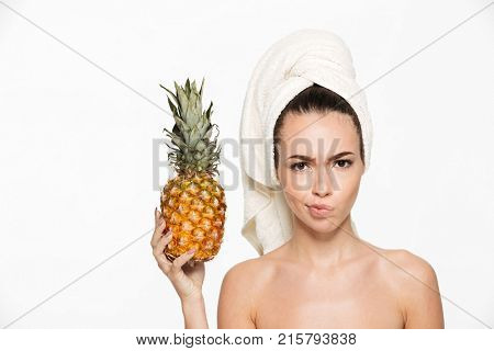Beauty portrait of an upset unsatisfied half naked woman with a towel wrapped around her face holding pineapple and looking at camera isolated over white background