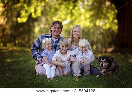 A portrait of a family of 5 blonde haired caucasian people sitting outside under the trees with their adopted pet German Shepherd Mix Breed Dog.
