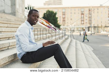 Happy black businessman working on laptop. African-american insuranse agent checking email on computer, sitting on the stairs outdoors in urban area