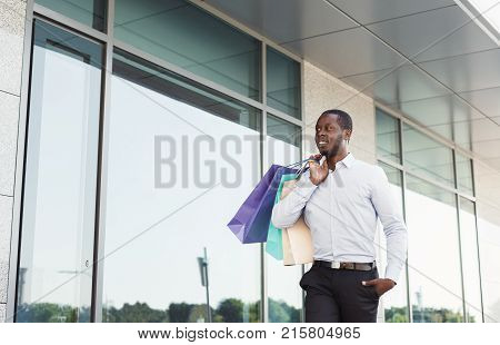 Happy black man with shopping bags walking along modern boutiques street. Successful business and shopping concept