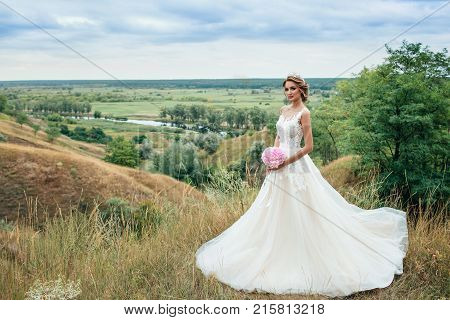 Beautiful Young Bride Girl Smiling, Standing In Wedding Dress. Overlooking The Beautiful Green Valle