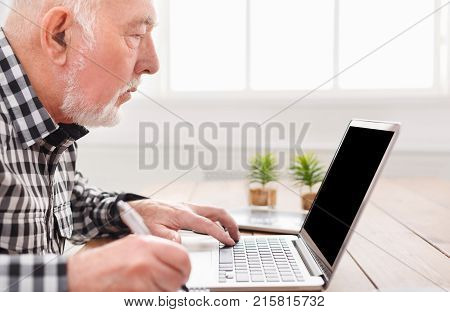 Mature man using laptop and writing in notepad at home desk. Education, recipe concept