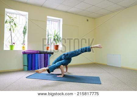 Young brunette of American origin sits on yoga mat and performs longitudinal twine. Short-haired sportswoman dressed in blue sports pants and blue t-shirt. Room light and spacious, under wall multi-colored yoga mats. Concept of to maintain body in good ph